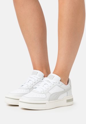 White leather sneakers with gray accents, perforated toe box, round laces, and elevated rubber sole. Minimalist design with a sporty aesthetic.