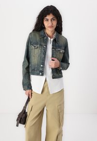 Giacca di denim blu scuro con finitura sbiadita, tasche frontali, bottoni metallici e design cropped, indossata sopra una blusa bianca e pantaloni beige.