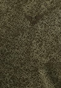 Tissu vert foncé avec un motif texturé de lignes entrecroisées. Un emblème brodé avec des ailes est placé près du haut.