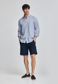 Jasnoniebieska lniana koszula z guzikami, granatowe szorty i ciemne buty wsuwane. Koszula ma luźny krój z podwiniętymi rękawami i małym logo.