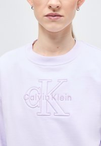 Γυναίκα που φοράει ανοιχτό μωβ φούτερ Calvin Klein με κεντημένο λογότυπο CK και μικρούς κρίκους σκουλαρίκια, εμφανιζόμενη από τον λαιμό έως το άνω μέρος του στήθους.