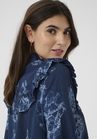 Mörkblå blus med blommönster och volanger vid axlarna, med en mjuk textur och ljusblå blommönster mot en mörk bakgrund.