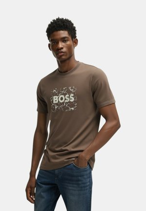 Camiseta de manga corta marrón con un logo blanco de "BOSS" sobre un fondo rectangular texturizado, combinada con jeans de mezclilla azul.