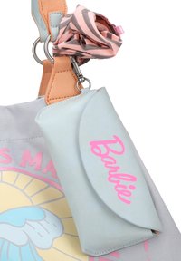 Lichtblauwe portemonnee met roze "Barbie" logo. Zacht materiaal, design met afgeronde klep, bevestigd aan een handtas met een roze en grijze gestreepte scrunchie.