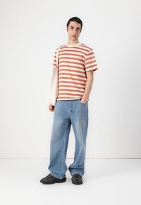 Striped t-shirt in coral and cream, paired with light blue wide-leg jeans. Model holds a canvas tote and wears black sneakers.