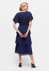 Vestido azul marino con mangas cortas acampanadas, diseño en capas y detalles de cuentas. La tela texturizada presenta patrones ondulados. Se ven zapatos de tacón alto.