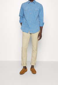 Light blue button-up shirt with two chest pockets, beige rolled-up chinos, and brown suede loafers. Smooth fabric, casual fit, tailored design.