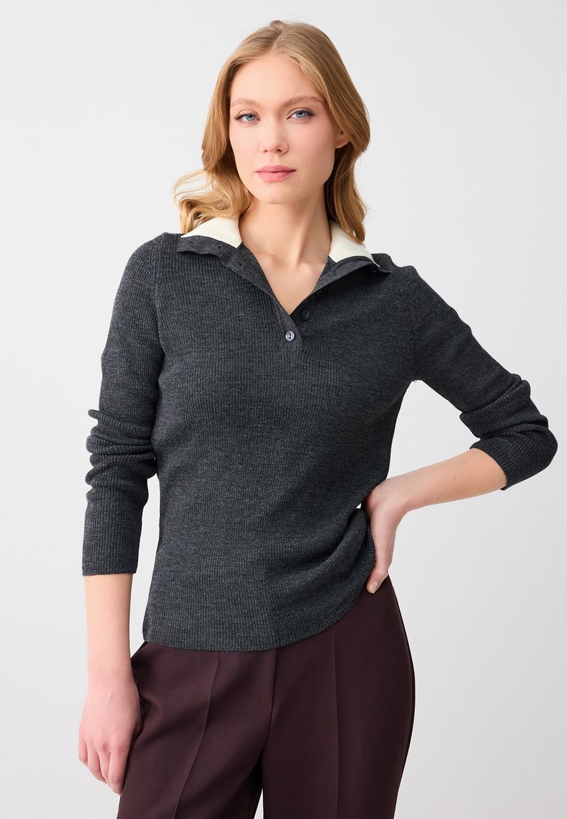 Femme blonde aux cheveux ondulés portant un pull en maille gris foncé avec un col blanc, posant avec une main sur la hanche, fond neutre.