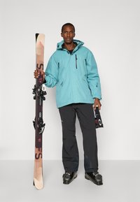 Türkise Skijacke mit Kapuze, schwarze Skihosen, schwarze Skischuhe, hält Skier mit Bindungen. Schlichter Hintergrund.