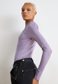 Langermet lilla treningsgenser med slim fit, med en logo på brystet og glatt, elastisk stoff. Kombinert med mørke denimbukser.