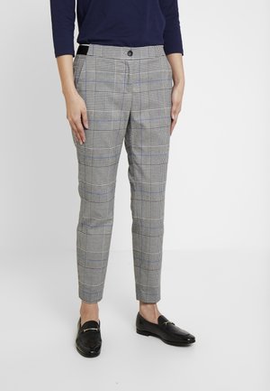 Person iført grå ternede skræddersyede ankellange bukser med en marineblå top og sorte loafers, stående foran en hvid baggrund.