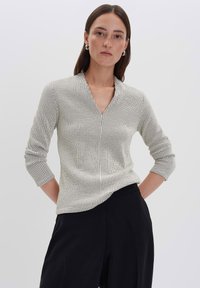 Una donna con capelli castani lisci indossa una maglietta bianca a maniche lunghe con scollo a V e un motivo a griglia nera, insieme a pantaloni neri a gamba larga, in piedi con le mani dietro la schiena.