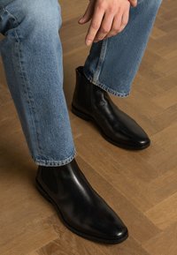 Stivali Chelsea in pelle nera con pannelli laterali elasticizzati, punta affilata e texture liscia, abbinati a jeans in denim blu.