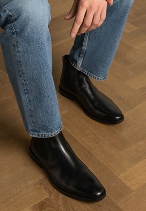 Bottines Chelsea en cuir noir avec des panneaux latéraux élastiques, forme de pointe aigüe et texture lisse, associées à un jean en denim bleu.