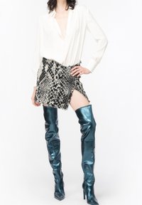 Vit blus med djup V-ringning, svart och vit mönstrad minikjol med fransar, och blanka turkosa överknästövlar.