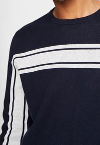 Navyblaues Strickpullover mit zwei weißen Streifen, die vertikal entlang der Ärmel verlaufen, einem gerippten Rundhalsausschnitt und einer strukturierten Stoffoberfläche.