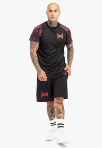 Sort atletisk t-shirt med lilla og røde mønstrede korte ærmer, parret med sorte shorts med røde "Tapout" logoer. Hvide sneakers og sokker.