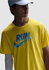 Camiseta deportiva amarilla con un logo azul de Nike que presenta la palabra "RUN", de manga corta, cuello redondo y textura que absorbe la humedad.