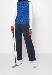 Person i mönstrad blå zip-up väst och marinblå byxor som håller en golfklubba och står framför en enkel vit bakgrund.