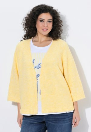 Donna con capelli ricci e scuri che indossa un cardigan giallo chiaro aperto sul davanti, una T-shirt bianca con stampa grafica e jeans blu, in piedi davanti a uno sfondo bianco.