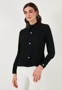 Black long-sleeve blouse with a classic collar, embellished buttons, and a smooth texture, paired with white pants for contrast.