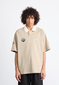 Joven con cabello rizado, vistiendo una camiseta tipo polo beige con logos y pantalones negros, de pie contra un fondo claro y liso.