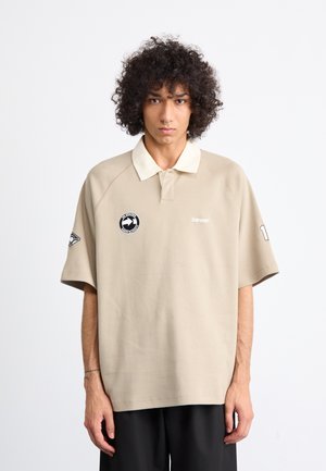 Young person with curly hair wearing a beige polo shirt with logos and black pants, standing against a plain light background.