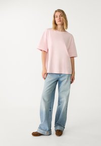 Roze oversized t-shirt met korte mouwen, gecombineerd met lichtblauwe wijde jeans. Zachte textuur en eenvoudig ontwerp. Bruine moccasinschoenen.