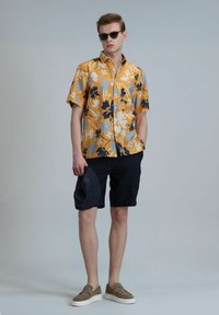 Korte mouwen gele bloemenoverhemd met blauwe en zwarte patronen, gecombineerd met donkere shorts en tan instap schoenen. Houdt een marineblauwe hoed vast.