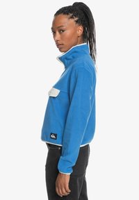 Blue fleece pullover with high collar and white accents, featuring a front pocket and a logo patch at the hem. Smooth texture.
