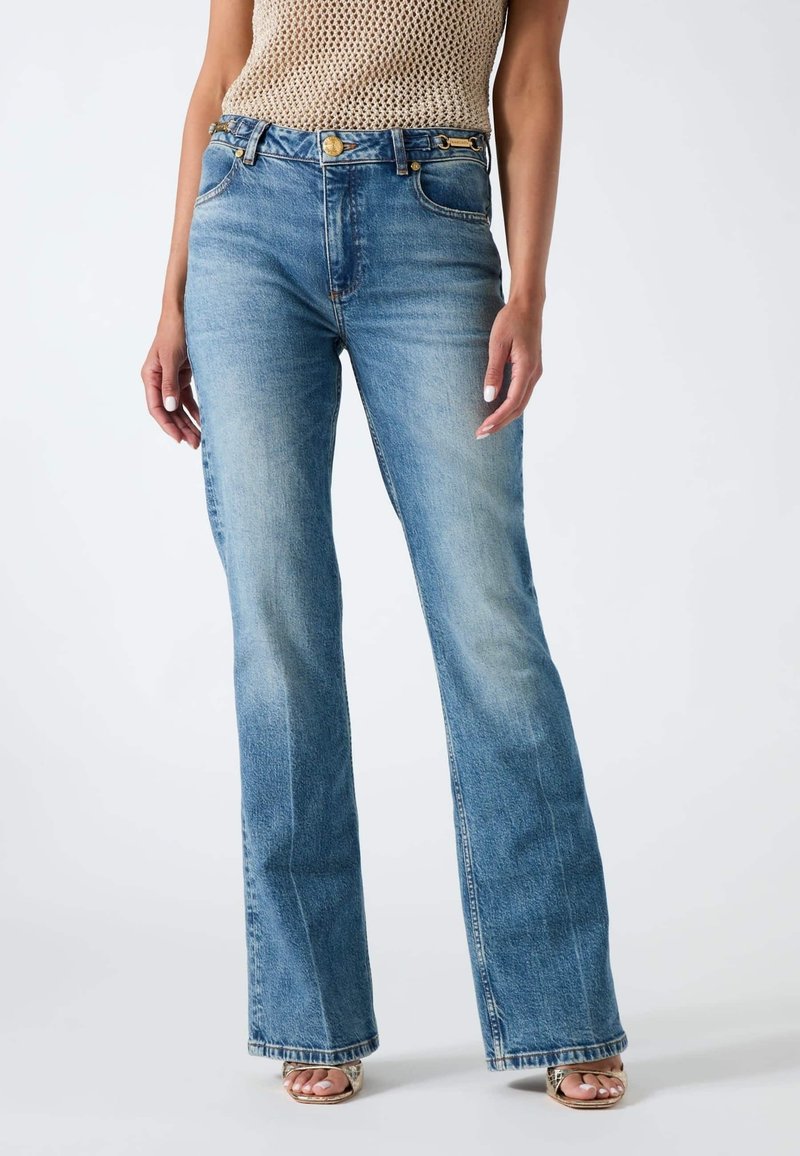 Femme portant un jean flare bleu clair taille haute, un top beige tricoté sans manches et des sandales à talons ouverts.