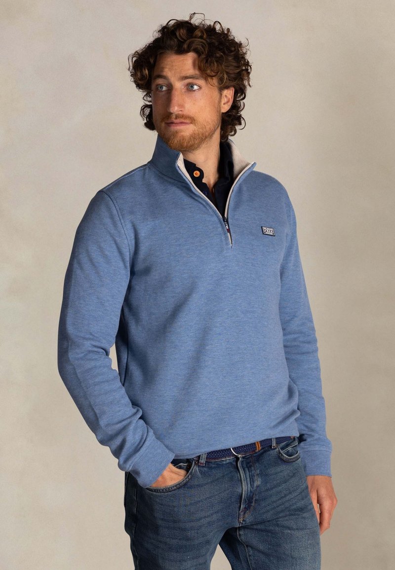Blauwe pullover met rits en lange mouwen, gemaakt van zacht materiaal, met een contrasterende kraag en een logo-patch op de borst. Gekocht in combinatie met een spijkerbroek.