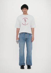 Hvit oversized t-skjorte med rød grafisk tekst, parret med lyseblå rette jeans og svarte sko, holder en sølvfarget gjenstand.