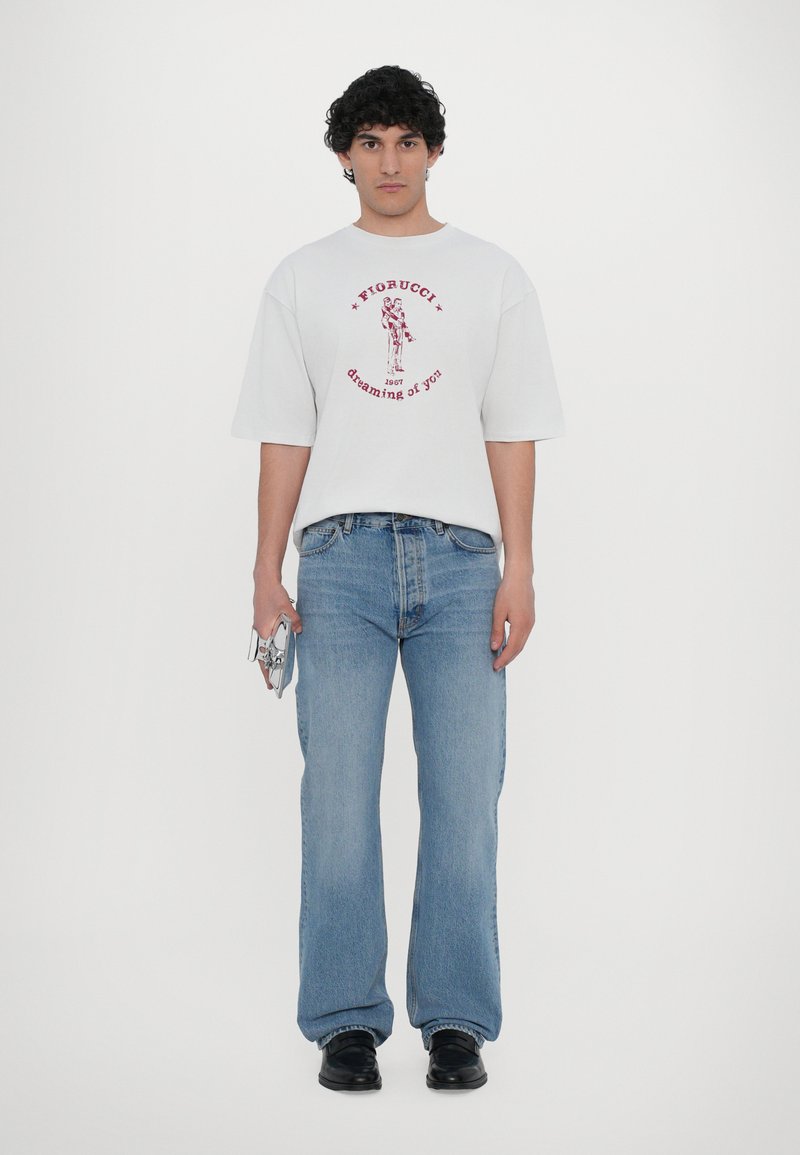 Hvit oversized t-skjorte med rød grafisk tekst, parret med lyseblå rette jeans og svarte sko, holder en sølvfarget gjenstand.