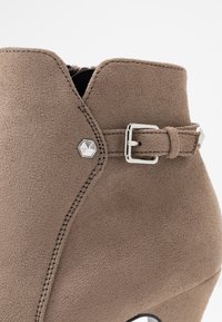 Gros plan sur une botte cheville en suède taupe avec un clou hexagonal argenté et un détail de boucle sur le côté.