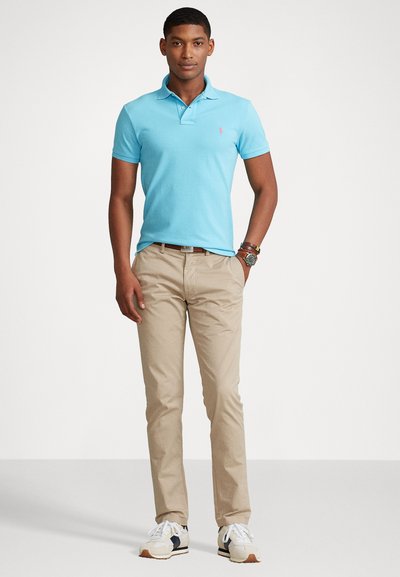 Ανοιχτό μπλε polo με γιακά, μπεζ chinos και λευκά αθλητικά παπούτσια με ναυτικές λεπτομέρειες. Ξεχωριστό ροζ λογότυπο πάνω στο μπλουζάκι.