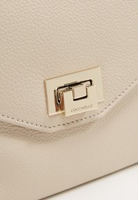 Vaaleanbeige nahkalaukku, jossa on teksturoitu pinta, kultasävyinen solki ja kohokuvioitu brändäys. Kulmikas muotoilu siisteillä ompeleilla.