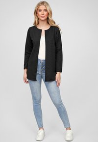 Zwarte textuurjas met een ronde halslijn en open voorkant, gedragen over een witte top en lichtblauwe skinny jeans, gecombineerd met witte sneakers.