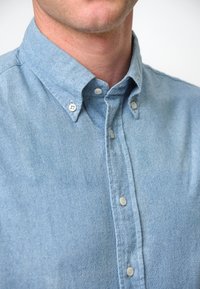 Camicia di jeans azzurro chiaro con colletto a bottone, caratterizzata da bottoni bianchi e un tessuto testurizzato. Nessun motivo o design visibile.
