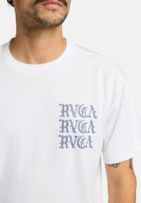 Witte katoenen t-shirt met een gestructureerde blauwe print op de linkerborst met herhaaldelijk "RVCA" logo in een gestileerd lettertype.