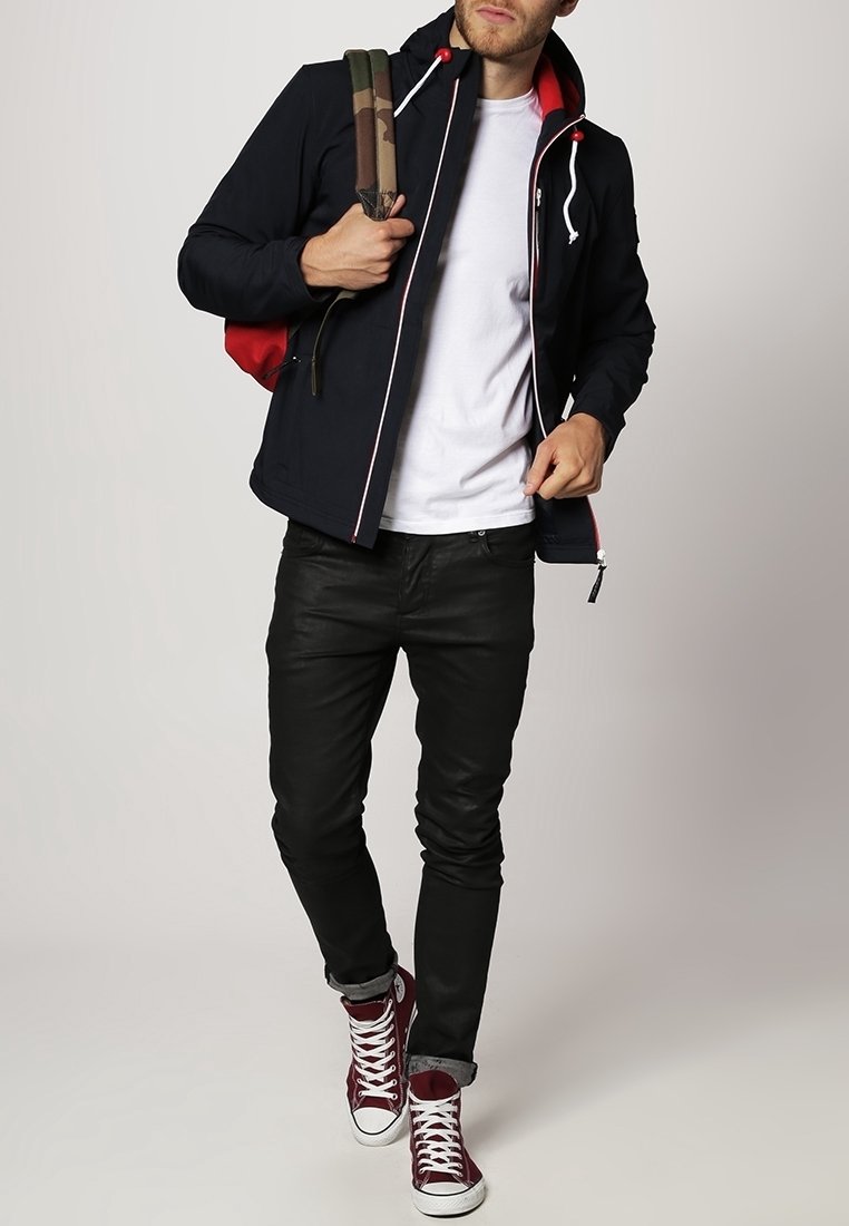 Homme portant une veste noire sur un t-shirt blanc, pantalon noir, baskets montantes bordeaux, portant un sac à dos camouflage sur une épaule.