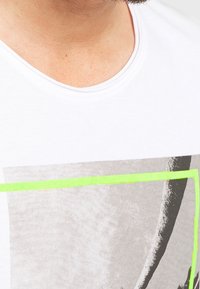 Hvid T-shirt med rund hals, prydet med et grå grafisk print omgivet af et klart grønt rektangel. Blødt, glat stof.