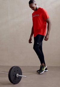 Nike Pro crvena majica kratkih rukava, crne kompresijske tajice i zelene/bijele atletske cipele, s bučicom na podu u blizini.