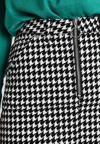 Gonna a stampa houndstooth bianco e nero con zip frontale e hardware in metallo, realizzata in tessuto testurizzato. Parte superiore verde parzialmente visibile.