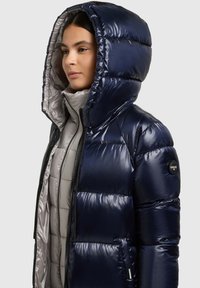 Tengerészkék pufi dzseki fényes kivitelben, kapucnival és szürke béléssel. Tűzött textúrával és oldalzsebbel rendelkezik. Cipzáras záródás.