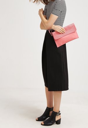Woman wearing black culottes, black open-toe ankle boots, striped top, holding pink clutch purse against plain background.