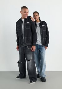 Zwei Modelle tragen passende schwarze Arbeitsjacken mit weißen Nähten und mehreren Taschen. Eine trägt abgewetzte graue Jeans; die andere hellblaue Jeans.