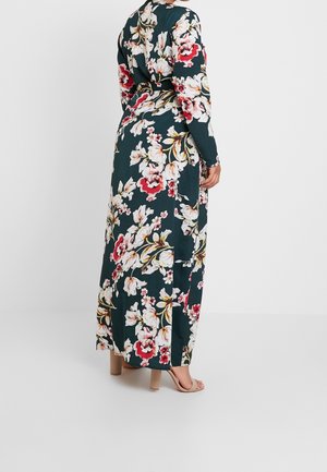 Vrouw staand, gekleed in een lange bloemenjurk donker groen met rode, witte en gele bloemen, gecombineerd met beige open-teen blokhaksandalen.