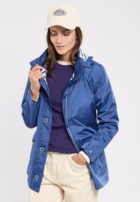 Giacca leggera blu con zip frontale, cappuccio e dettagli in bottone; foderato internamente con un motivo bianco e blu; abbinata a un maglione viola.