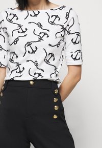 Biała bluzka z czarnym wzorem kotwicy i liny, okrągły dekolt i krótkie rękawy; w zestawieniu z czarnymi spodniami z złotymi guzikami.